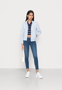 Casaco bomber azul claro, polo curta listrada em azul marinho, jeans ajustados azuis com costuras nos joelhos e ténis plataformas brancos. Pequena bolsa branca.