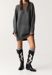 Pull en tricot gris avec un col rond et des poignets côtelés, associé à des bottes en cuir noir hautes jusqu'au genou ornées de motifs décoratifs blancs.