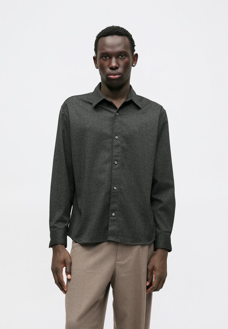 Chemise à manches longues en charbon avec un motif texturé, boutons contrastés et col classique, associée à un pantalon marron clair.