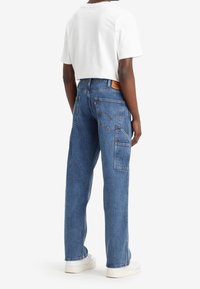 Persoon in blauwe rechte denim jeans met meerdere achterzakken en een witte cropped t-shirt, staande met de rug naar de camera.