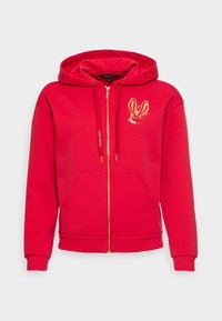 Sweat à capuche rouge à fermeture éclair fabriqué en tissu doux, avec une poche avant et des accents dorés, y compris un motif distinctif sur la poitrine.