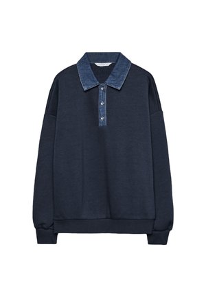 Vrouw draagt een marineblauwe trui, grijze shorts met krijtstreep, zwarte kniehoge laarzen en draagt een kleine zwarte schoudertas.
