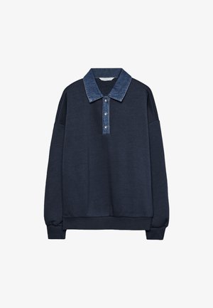 Vrouw draagt een marineblauwe trui, grijze shorts met krijtstreep, zwarte kniehoge laarzen en draagt een kleine zwarte schoudertas.