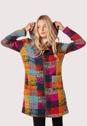 Donna che sistema il cappuccio su un lungo cappotto patchwork multicolore con zip e tasche anteriori, indossando pantaloni neri e una maglietta chiara sotto.