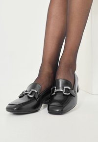 Svarta läderloafers med fyrkantig tå, silverdetaljer, låg klack och slät yta. Bar med genomskinliga svarta strumpor.