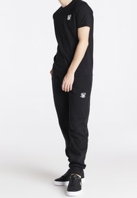 Zwarte t-shirt met korte mouwen en een klein wit logo op de borst, gecombineerd met zwarte sweatpants met een bijpassend logo. Zwarte schoenen met witte zolen.