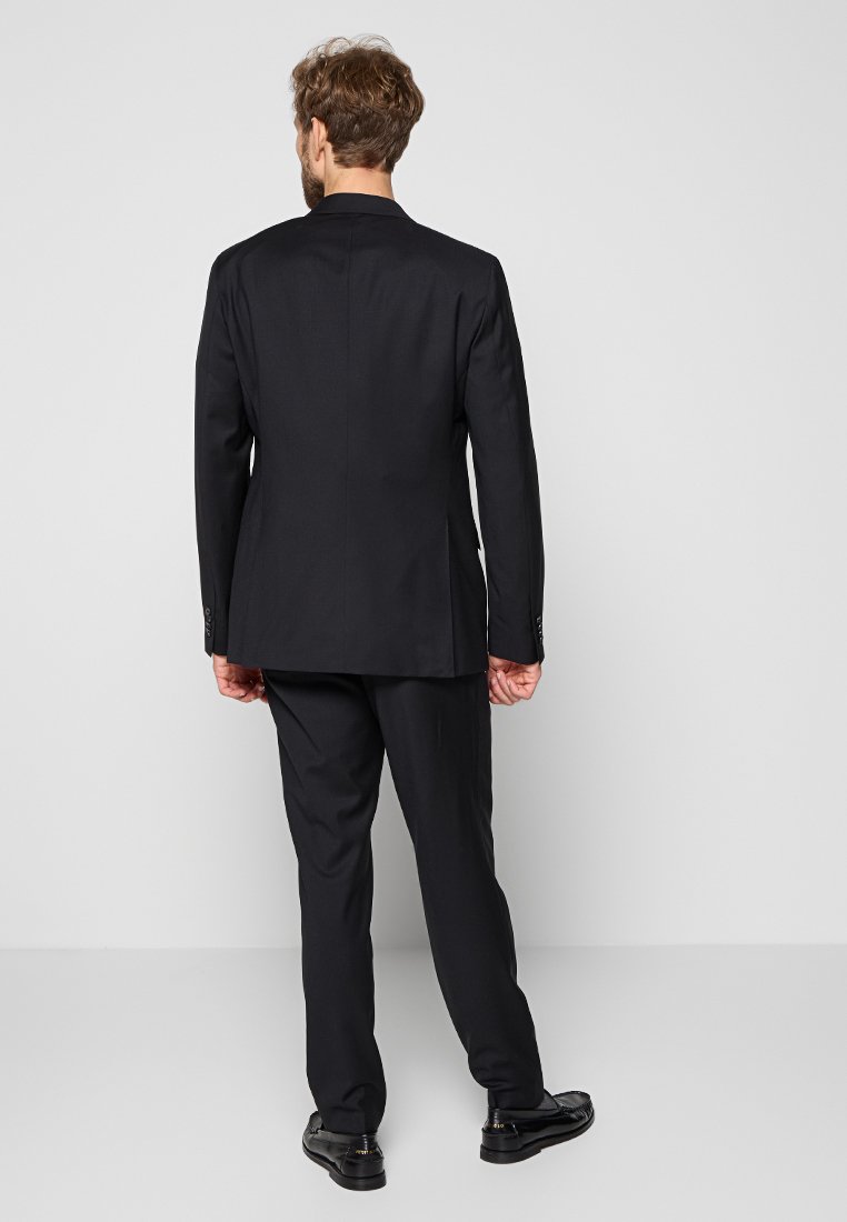 Man staat met de rug naar de camera in een strakke zwarte pak met een jas en broek, gecombineerd met zwarte nette schoenen, tegen een effen grijze achtergrond.