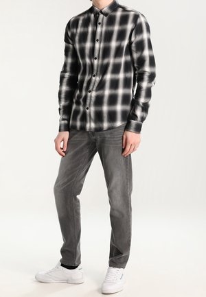 Camicia a maniche lunghe a scacchi bianco e nero con chiusura a bottoni, abbinata a jeans grigi e sneakers bianche, creando un look casual.