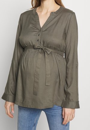 Vrouw draagt een olijfgroene zwangerschapsblouse met lange mouwen en een voorstrik, en blauwe jeans, afgebeeld van nek tot heupen.