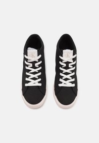 Svarta kanvas sneakers med vita snören, rund tå och texturerad gummisula. Logotypetikett på tungan i ljus färg, minimalistisk design.