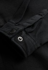 Polsino di giacca in denim nero con bottone a pressione e tessuto nero, con finitura liscia e dettagli di cucitura visibili.