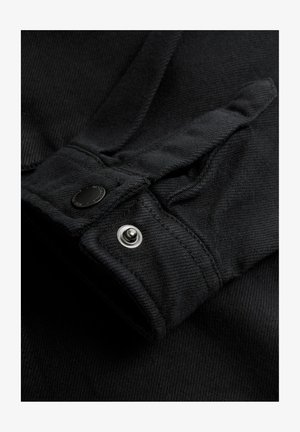 Fekete farmerruha kabátmandzsettája patenttal és fekete anyagtextúrával, sima felülettel és látható varrási részletekkel.