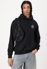 Black hooded sweatshirt with a front pocket and white graphic, paired with light blue denim jeans and a black crossbody bag.