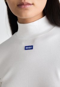 Bílý žebrovaný rolák s modrou obdélníkovou visačkou s nápisem "HUGO" bílými písmeny, označující značku. Hladká textura.