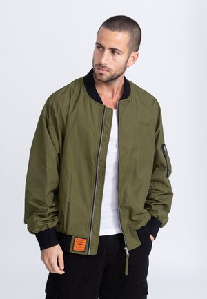 Giacca bomber verde oliva con zip frontale, polsini a coste, tasche laterali e una toppa sull'orlo inferiore. Tessuto morbido e vestibilità comoda.