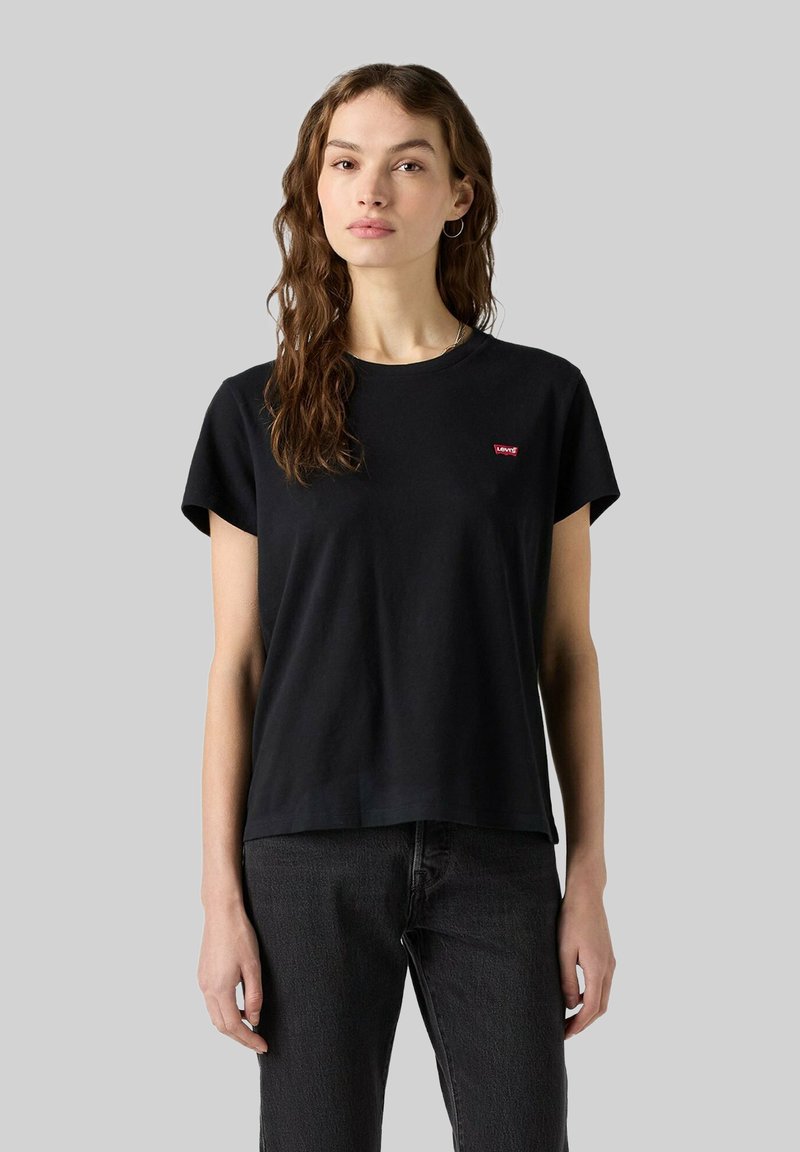 Junge Frau mit langen braunen Haaren, die ein schwarzes Levi's T-Shirt und dunkle Jeans vor einem grauen Hintergrund trägt.