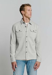 Lichtgrijze button-up shirt met twee borstzakken, zwarte knopen en een kraag. Gekozen over een wit shirt, gecombineerd met blauwe jeans.
