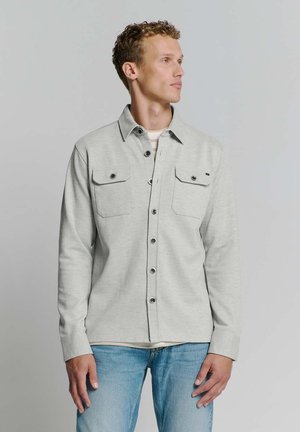 Hellgraues Hemd mit Knöpfen und zwei Brusttaschen, schwarzen Knöpfen und einem Kragen. Getragen über einem weißen Hemd, kombiniert mit blauen Jeans.