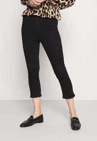 Černé zkrácené džíny s přiléhavým střihem a frayed lemem, doplněné černými mokasíny. Modelka má na sobě vzorovanou blůzu s volánkovým výstřihem.