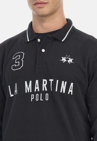 Svart bomullspolo med vit brodyr. Har en krage, tre-knapps placket och logotyp med två polospelare. Nummer "3" på vänster bröst.