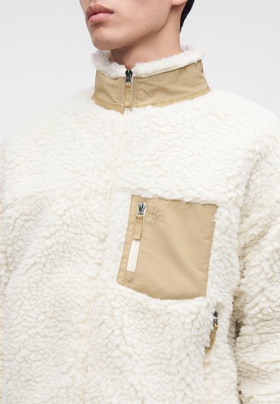 Κοντινό πλάνο ενός ατόμου που φοράει λευκό sherpa fleece μπουφάν με ταμπά γιακά και τσέπη στο στήθος με φερμουάρ.