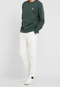 Zielona bluza z logo na piersi, w połączeniu z białymi, luźnymi spodniami i białymi sneakersami. Prosty, casualowy design.