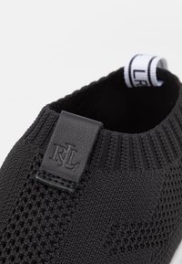 Chaussure noire en maille à enfiler avec un motif texturé, languette en cuir embossée « RLL » et boucle de talon en tissu blanc avec texte noir.