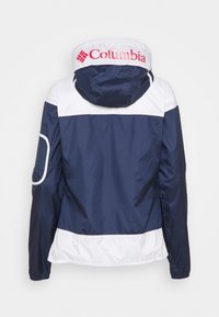 Coupe-vent Columbia bleu marine et blanc avec capuche, logo rouge sur la capuche, manches longues et détail en mesh sur la manche gauche.