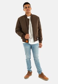 Brun bomberjacka med ribbade muddar, ficka med dragkedja, bärs över en vit skjorta, ihop med ljusblå jeans och bruna snörstövlar.