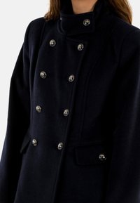 Cappotto di lana blu navy con colletto alto, design doppiopetto e dettagli in bottoni argentati. Presenta tasche laterali e una texture morbida.