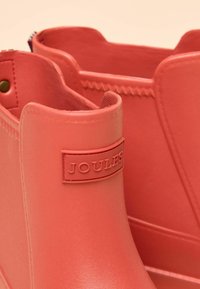 Gli stivaletti in gomma color corallo presentano una texture liscia e un logo Joules in rilievo sul lato; i dettagli includono cuciture e una fodera interna decorativa.