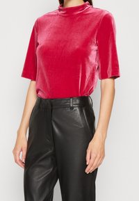 Haut ajusté à manches courtes en velours rouge plush avec un col montant ; associé à un pantalon en cuir noir taille haute avec des poches latérales.