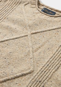 Beiger gestrickter Pullover mit strukturierten diagonalen und vertikalen Mustern sowie einem dunkelblauen Etikett mit dem Markennamen in der Nähe des Kragens.