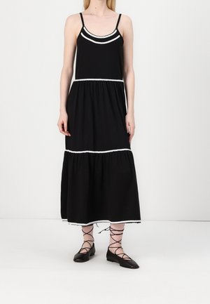 Woman wearing a sleeveless black midi dress with white trim and black lace-up ballet flats standing against a plain background.