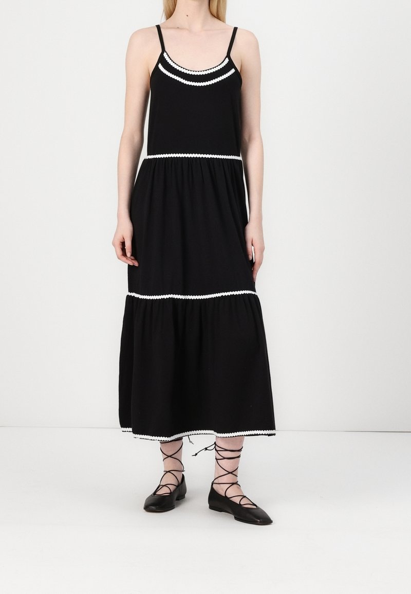 Femme portant une robe midi noire sans manches avec un bord blanc et des ballerines noires à lacets, debout devant un fond uni.