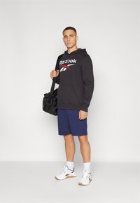 Crna hoodie s Reebok logotipom, tamnoplave kratke hlače, bijele tenisice i crna torba. Glatka tkanina, opušteni kroj, jednostavan dizajn.