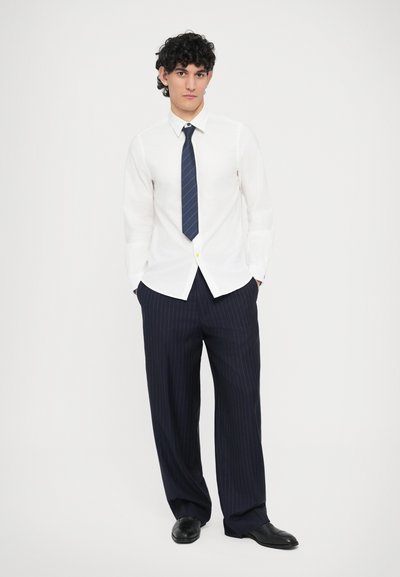 Camicia bianca a bottoni, pantaloni a righe blu navy, scarpe nere; presenta una cravatta stretta a righe diagonali, vestibilità comoda e maniche lunghe.