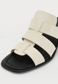 Sandales à lanières avec trois larges sangles en synthétique crème, une semelle noire et un design à bout carré, avec un accent argenté sur les lanières.