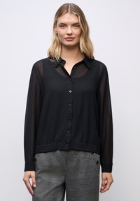 Sort transparent bluse med knaplukning foran, krave og elastisk talje. Kombineret med grå rute-mønstrede cropped bukser med knapdetaljer.