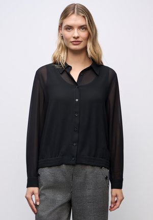 Schwarze durchsichtige Bluse mit Knopfleiste, Kragen und elastischem Taillenbund. Kombiniert mit grauen karierten cropped Hosen mit Knopfdetails.