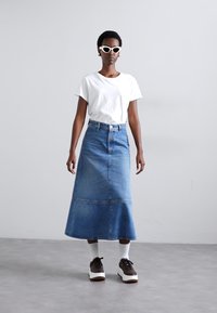 Denim maxi-hame, jossa on liehuva muoto, sininen pesu, ommellut yksityiskohdat ja takatasku. Yhdistetty valkoisen T-paidan ja korkokenkien kanssa.