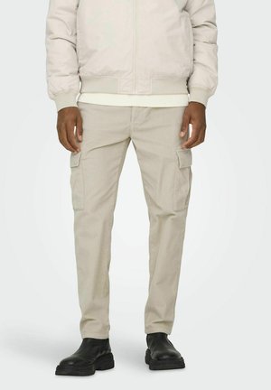 Pantalones cargo beige con una textura acanalada, que presentan dos bolsillos laterales, un corte recto, combinados con botas negras hasta el tobillo y una chaqueta bomber clara.