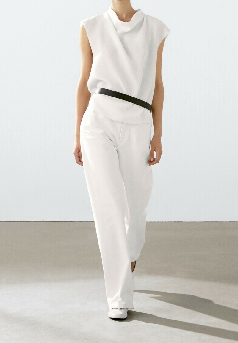 Personne marchant en haut blanc sans manches et pantalon blanc ample avec une ceinture noire et des chaussures blanches sur un fond uni.