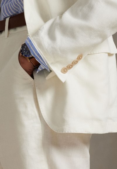 Lininis švarkas kreminės baltumo spalvos su keturiais smėlio spalvos sagomis ant rankogalių. Matoma mėlynomis juostelėmis marginta marškinėliai ir apvalus laikrodis su tamsiu ciferblatu ir auksinėmis detalėmis.