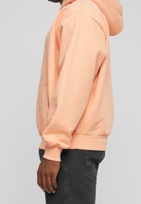 Persona che indossa una felpa color pesca con tasca frontale e jeans grigio scuro, mostrata di lato su uno sfondo semplice.