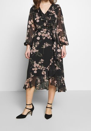 Frau trägt ein schwarzes Midikleid mit Blumenmuster, Rüschen, langen transparenten Ärmeln, einem gebundenen Taillengürtel und schwarzen Absatzschuhen mit Knöchelriemen.