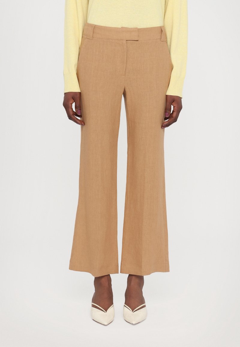 Femme portant un pantalon beige à jambes larges et des chaussures crème à bouts pointus, debout devant un fond clair uni.
