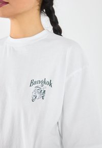 Balta medvilnės marškinėliai su žalia tuk-tuk grafika ir žodžiu "Bangkok" stilingu šriftu šalia kairės krūtinės.