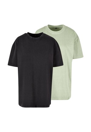 Femme aux cheveux bouclés portant un t-shirt ample gris foncé à manches courtes, debout devant un fond gris uni.