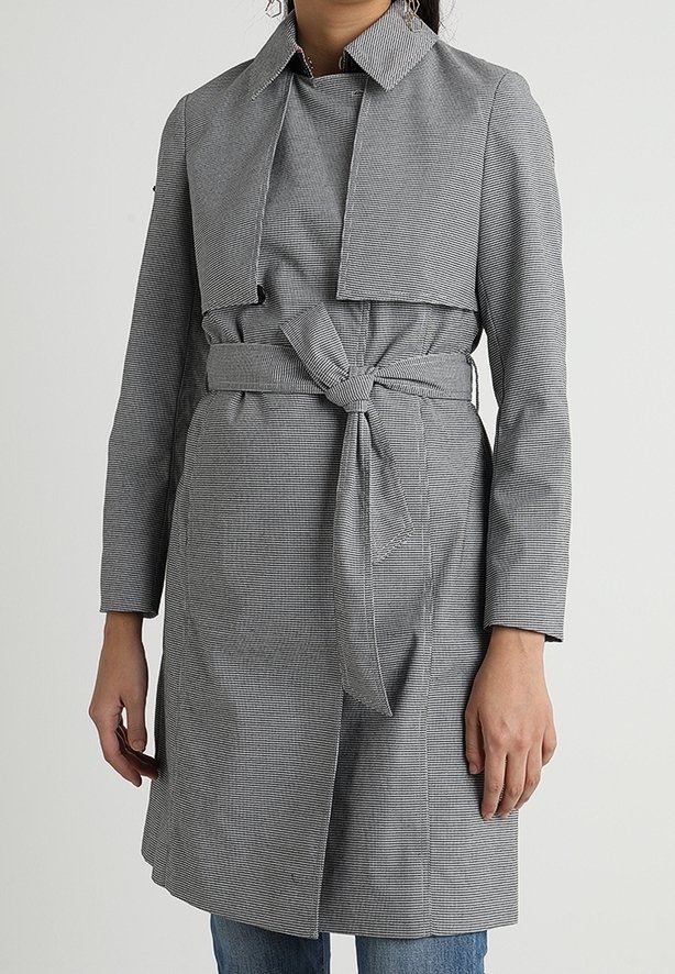 Femme portant un trench-coat gris jusqu’aux genoux, avec une ceinture nouée en nœud à l’avant, des manches longues et un large col, debout devant un fond uni.